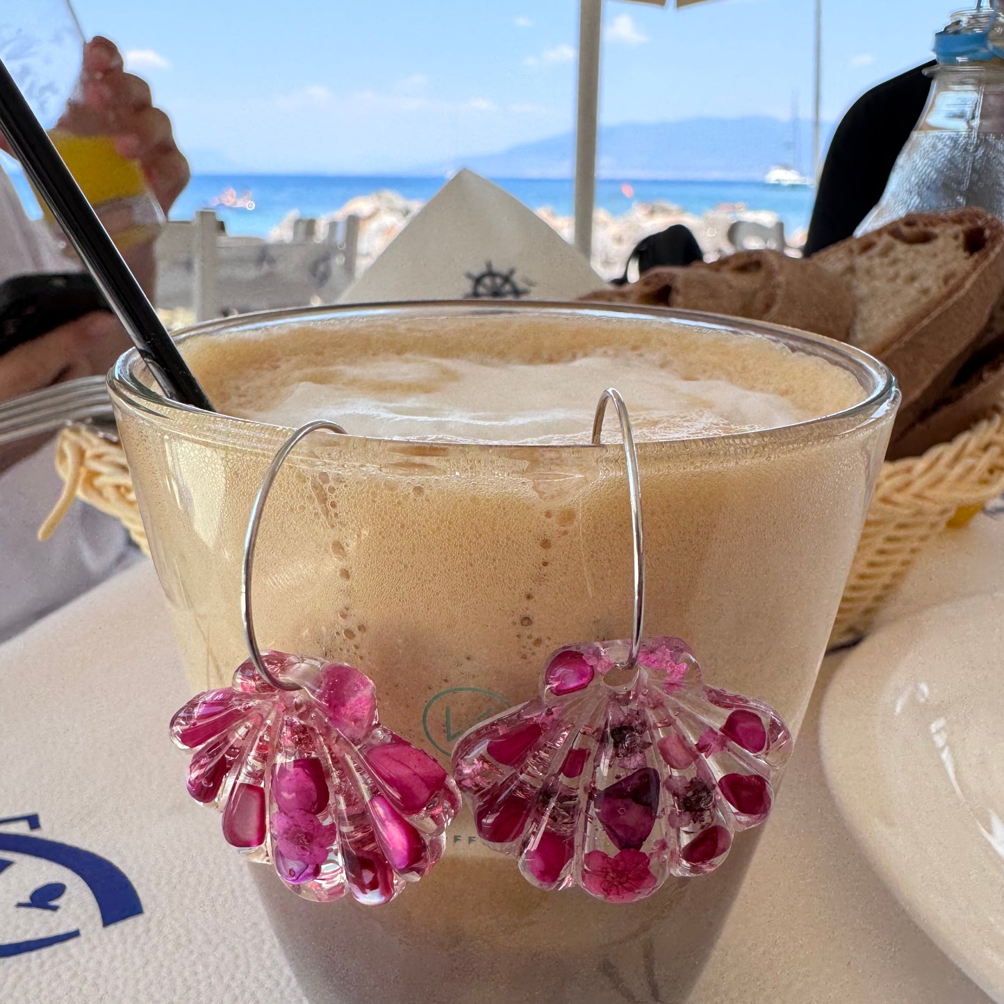 A close-up of a frothy light brown beverage in a clear glass, decorated with two pink flower-shaped garnishes and a metal ring, set on a white tablecloth with blue graphics.