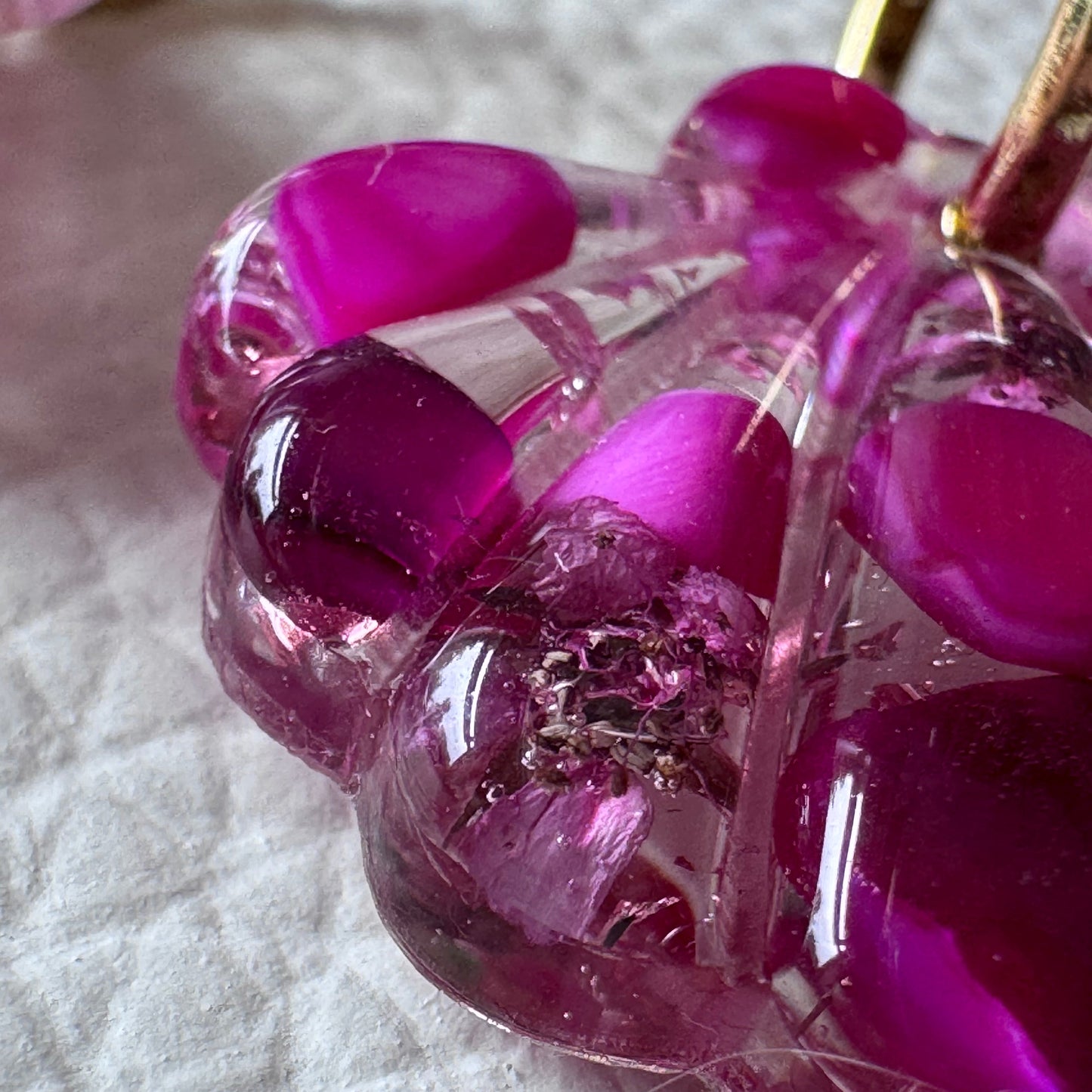 A close-up of a translucent pink and magenta resin charm or accessory with organic bubble shapes and a small metallic gold-toned element, resting on a white textured fabric background.