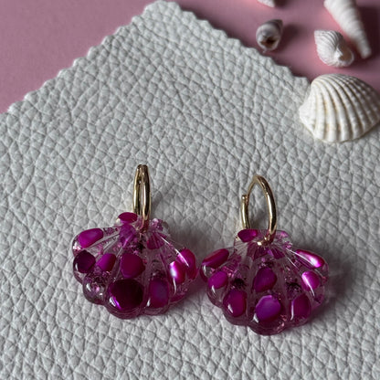 A pair of handmade magenta pink beaded dangle earrings with gold-toned hooks, displayed on a textured white surface next to seashells on a pink background.