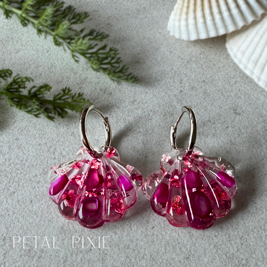 A pair of handmade seashell-shaped drop earrings in translucent pink and magenta resin with embedded glitter, displayed on a textured gray surface with decorative seashells and greenery.