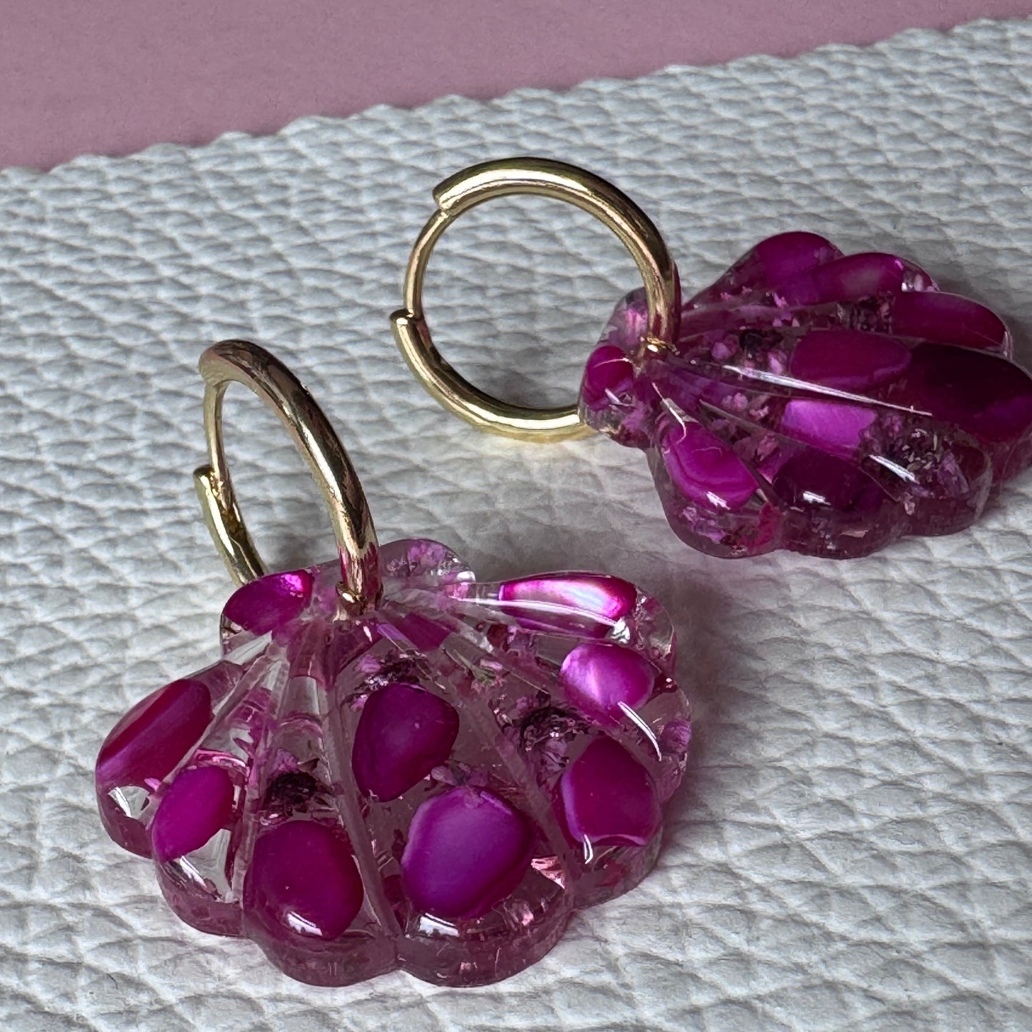 A pair of handmade earrings featuring translucent magenta pink flower-shaped charms with gold-tone hoops, displayed on a textured white surface.